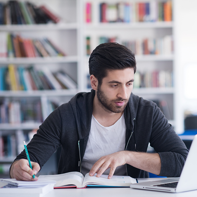 student at a desk studying from a book and laptop
