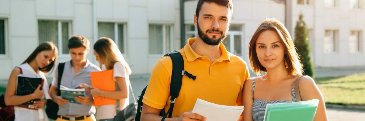Two happy students with backpacks and books in their hands smiling at camera while standing on background of university and friends