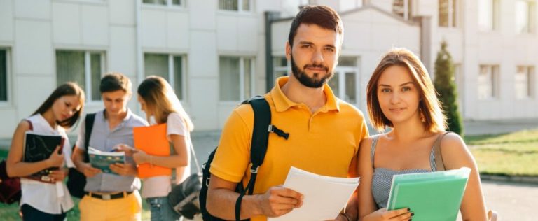 Two happy students with backpacks and books in their hands smiling at camera while standing on background of university and friends