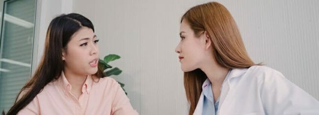 Doctor talking to unhappy teenage patient in exam room. Asian wo Doctor talking to unhappy teenage patient in exam room. Asian wo