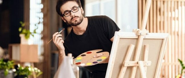 Freelancer man painting with brush standing behind easel. an painting with brush standing behind easel